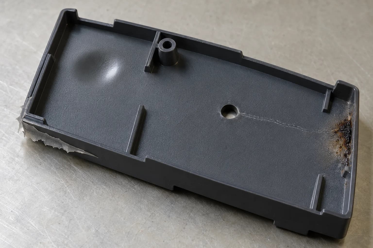 Un coperchio tecnico in plastica grigio scuro, visto dall'interno su un banco di controllo: un risucchio in alto a sinistra, una bava lungo il bordo a sinistra, una linea di giunzione che parte dal foro centrale e una bruciatura sull'angolo a destra.
